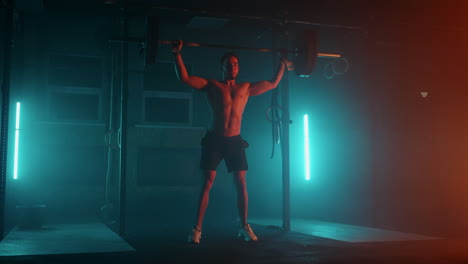 Männlicher-Powerlifter-In-Blau-orangefarbenem-Neonlicht-Bereitet-Sich-Auf-Ein-Training-Im-Fitnessstudio-Vor.-Ein-Kräftiger-Mann-Mit-Nacktem-Oberkörper-Macht-Beim-Heben-Einer-Langhantel-über-Den-Kopf-Eine-Ruckübung-Aus-Dem-Gewichtheben.